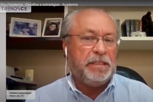 Imagem: Foto do reitor da UFC, Prof. Cândido Albuquerque, utilizando fones e falando na transmissão. Ao fundo, há porta-retratos e livros em uma prateleira na parede do escritório