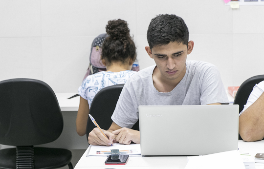 Imagem: Estudante sentado olhando para a tela de um notebook que está sobre uma mesa a sua frente; no fundo há uma estudante sentada de costas, estudando (Foto: Viktor Braga/UFC)