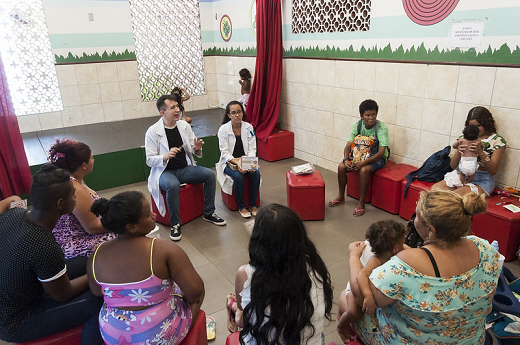 Imagem: foto de mulheres sentadas formando um círculo com duas pessoas de jaleco branco dando algumas instruções às demais