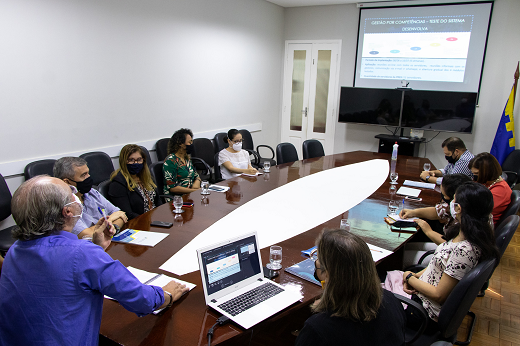 Imagem: foto de uma sala de reuniões com pessoas ao redor de uma mesa oval observando dados em um telão