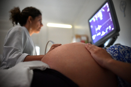 Imagem: Mulher grávida deitada; ao lado, uma profissional de saúde passa um instrumento sobre a barriga da mulher e acompanha as imagens que aparecem em um monitor (Foto: Agência Brasília)