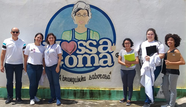 Imagem: seis pessoas posam para foto em frente a uma parede branca com desenho de uma camponesa com a frase Nossa Goma