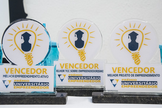 Imagem: Três troféus de uma edição passada do Prêmio Universitário Empreendedor sobre uma superfície plana (Foto: Viktor Braga)