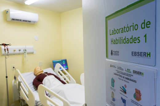 Imagem: Quarto de hospital; em primeiro plano há uma porta entreaberta; em segundo plano há um boneco simulando um paciente deitado em uma cama (Foto: Viktor Braga/UFC)