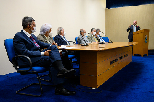 Imagem: foto de homens de paletó e gravata sentados em frente à uma mesa de madeira. Ao fundo, um homem branco, de pé, também de traje social, fala no púlpito