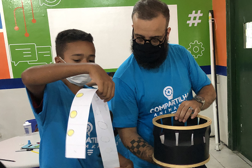 Imagem: criança negra de blusa azul segura uma fita de papel com desenhos. Ao seu lado, um jovem de blusa azul segura uma caixa preta enquanto observa os desenhos da criança. Os dois estão no centro de uma sala de aula. Ao fundo, há uma parede verde com lousa branca. 