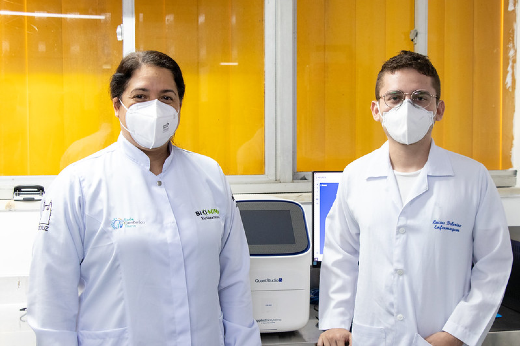 Imagem: Foto dos pesquisadores Veridiana Pessoa e Lucas Delerino, vestidos de jaleco e utilizando máscaras de proteção facial, no laboratório do HEMOCE