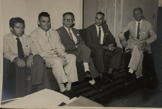 Imagem mostra foto preto e branco de cinco homens sentados, sendo quatro em um sofá do lado esquerdo e um em uma cadeira do lado direito 