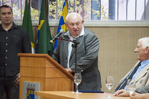 Imagem mostra professor Tarquínio discursando em um púlpito ao centro, com um homem em pé do lado esquerdo e outro sentando no lado direito acompanhando a fala 