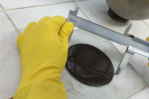 imagem mostra mistura em estado líquido de cor escuro sendo medida por parquímetro, que é manuseado por uma mão com luva amarela