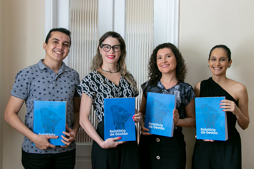 Imagem mostra quatro pessoas, sendo um homem à esquerda e três mulheres à direita, estão em pé, lado a lado, cada um segurando um exemplar do relatório, cuja capa predomina a cor azul 
