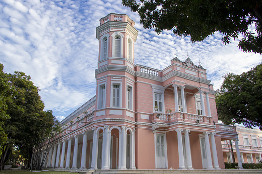 Imagem: Foto da fachada da reitoria. É um prédio de cor rosa com detalhes brancos nas colunas e janelas. Ao fundo um céu azul cheio de pequenas nuvens. Ao redor da reitoria o verde das mangueiras. Fim da descrição.