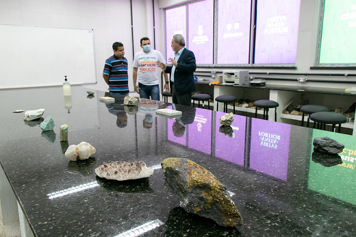 Imagem: Reitor conversa com professores no Laboratório de Geologia; na mesa, frente a eles, amostras de rochas