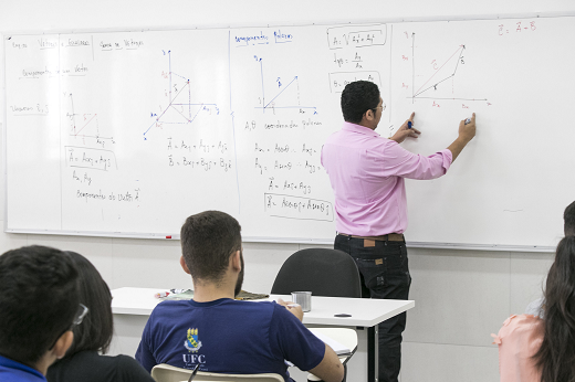 Imagem: professor de costas, em pé, de camisa rosa e calça preta, escrevendo em quadro branco, enquanto alunos assistem a aula sentado