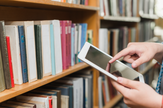 Imagem: Foto de uma pessoa segurando um tablet em frente de uma estante de livros