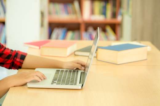 Imagem: mão de pessoa digitando no computador com livros ao fundo