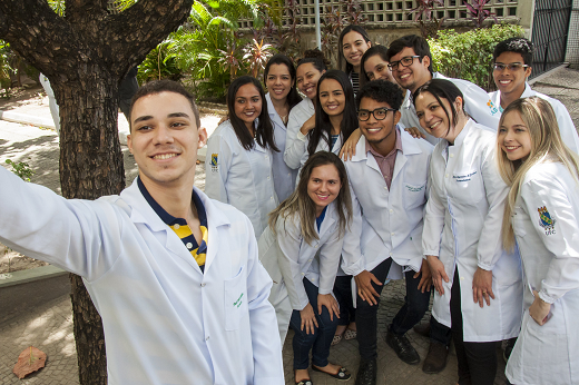 Imagem: Grupo de 13 estudantes de medicina, formado por homens e mulheres, posando para foto próximo a uma árvore; vestem o jaleco branco sobre a roupa (Foto: Viktor Braga/UFC)