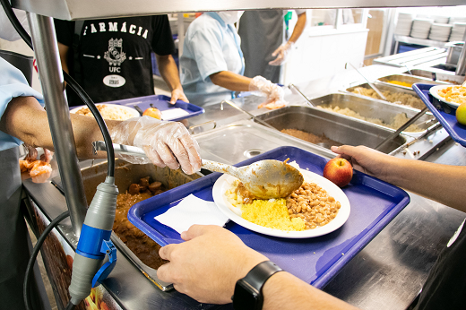 Imagem: Estudante segura uma bandeja com um prato de comida em um refeitório do Restaurante Universitário da UFC (Foto: Ribamar Neto/UFC)