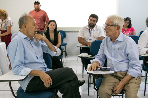 Imagem: dois homens brancos grisalhos estão sentados em um auditório, um de frente para o outro