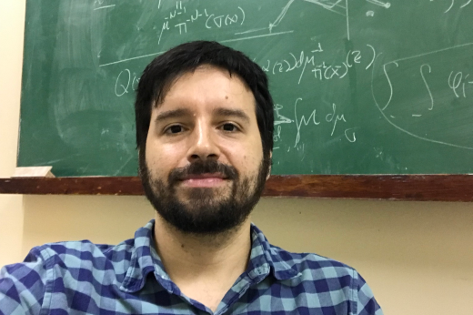 Imagem: Foto mostra o professor Mauricio Poletti à frente de uma lousa de sala de aula. O professor é um homem branco, cabelo preto, de barba, usa camisa xadrez azul  (Foto: Acervo pessoal)