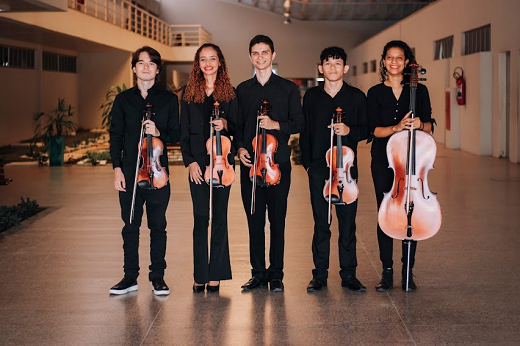 Imagem: cinco jovens posam para a foto segurando instrumentos de cordas. Todos estão em pé e de roupas pretas