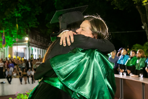Imagem: foto de duas mulheres jovens se abraçando. Uma está de beca e outra de veste talar preta com detalhes verdes