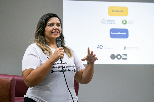 Imagem: foto de uma mulher branca, de cabelos loiros, falando ao microfone. Há uma projeção atrás dela