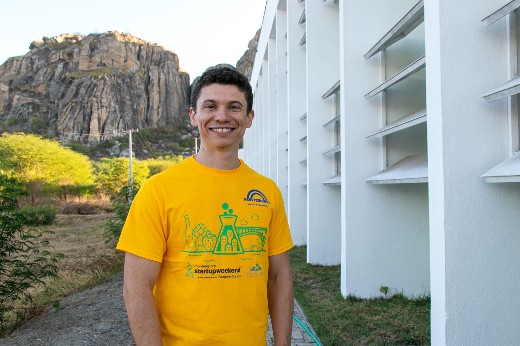 Leonel Júnior, egresso do campus, em pé, sorrindo para a foto, em uma área externa do campus; em segundo plano há um monólito típico da região de Quixadá (Foto: Viktor Braga/UFC)