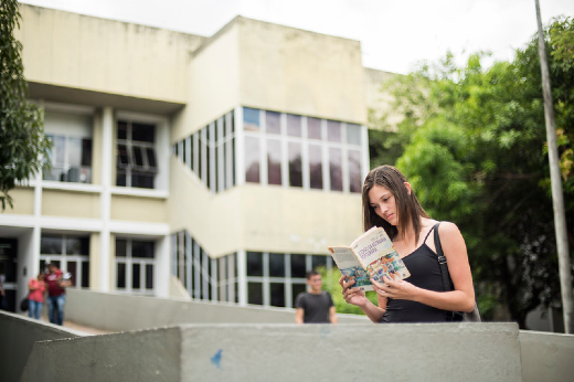 Imagem: Menina lê livro, em primeiro plano, e, ao fundo, aparece a Biblioteca Central do Campus do Pici
