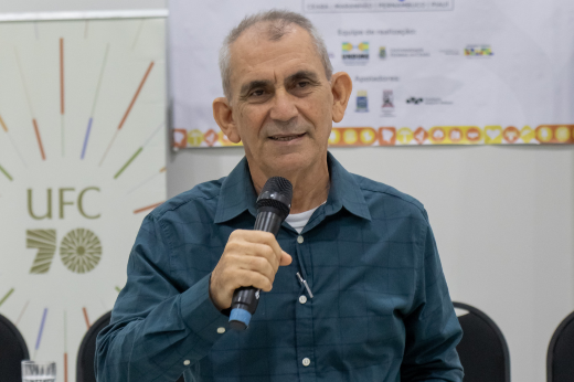 Imagem: foto do prof Manoel Andrade Neto, um homem de meia idade, com cabelos curtos grisalhos, usando camisa de botão na cor verde escura e falando ao microfone