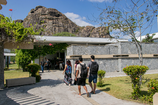 Imagem: foto do Campus de Quixadá, onde se vê pessoas de costas andando para entrar no bloco didático. Pode-se ver também uma grande rocha ao fundo e árvores ao redor 