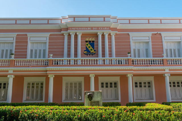 Imagem: fachada lateral da reitoria da UFC, um prédio com térreo e primeiro andar, em estilo clássico, nas cores salmão e branco (janelas, colunas, varanda), com um brasão centralidade instalado no primeiro andar e um jardim à frente