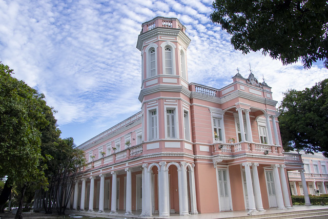 Foto da fachada da reitoria. É um prédio de cor rosa com detalhes brancos nas colunas e janelas. Ao fundo um céu azul cheio de pequenas nuvens. Ao redor da reitoria o verde das mangueiras. Fim da descrição.