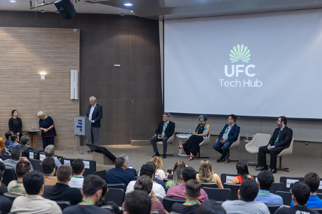 Imagem: Foto mostra a vista do palco de um grande auditório onde há pessoas sentadas nas cadeiras olhando para o palco, que tem representantes da UFC sentados e um telão atrás deles onde se pode ler Tech HUb UFC