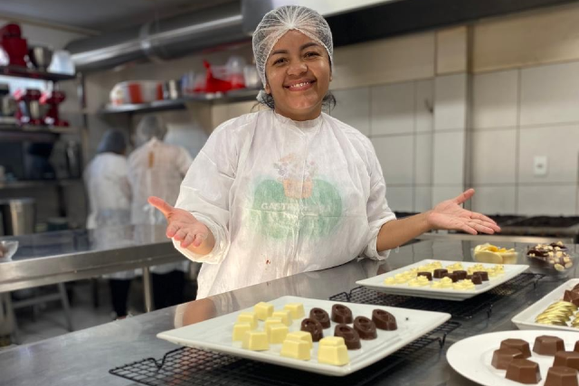 Imagem: foto de uma mulher negra com touca branca na cabeça. Ela está em frente a uma bancada mostrando chocolates. 