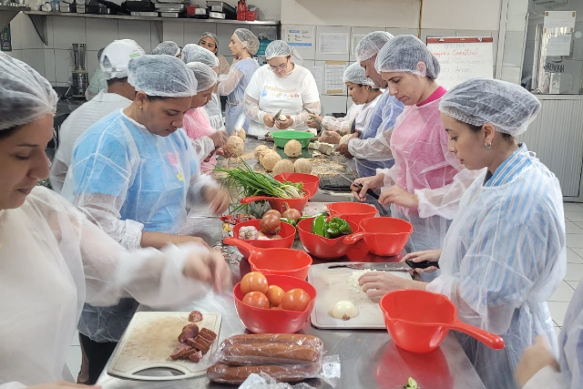 Imagem: Foto com mulheres ao redor de uma mesa retangular numa cozinha. Elas vestem touca e bata de tecido branco. Na mesa, há vários tipos de alimentos e utensílios de cozinha.