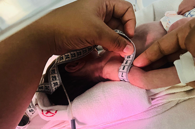 imagem: Mãos de um profissional de saúde medem, com fita métrica, o perímetro cefálico de um bebê recém-nascido. O bebê, de pele clara e cabelos escuros, está deitado sobre um tecido branco e parcialmente coberto por ele. Um dos braços do bebê está dobrado próximo ao rosto. A cena acontece em ambiente hospitalar, possivelmente em uma incubadora, com iluminação suave.