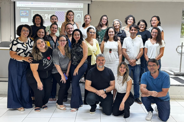 Imagem: várias pessoas posam para foto. Todas estão de calças e à frente de um telão de data show