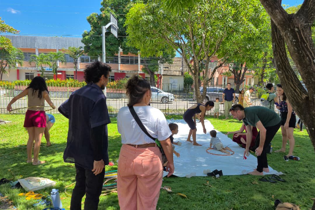 Imagem: foto de crianças brincando em um tapete estendido em um jardim. Adultos observam a cena