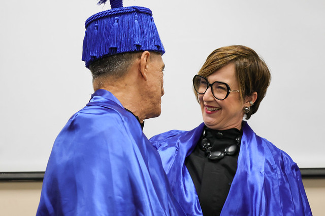 Imagem: Um homem idoso e uma mulher se cumprimentam sorrindo. Ambos vestem becas azuis; ele usa um barrete azul, e ela, óculos grandes e colar preto.