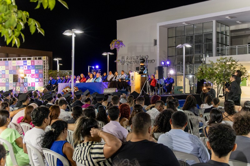 Imagem: Colação de grau da UFC em Sobral, realizada à noite. Formandos acompanham a cerimônia diante do palco, onde professores e autoridades acadêmicas participam da celebração.