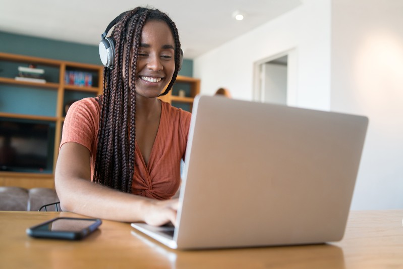 Imagem: Uma mulher negra, sorridente, usa fones de ouvido enquanto trabalha em um notebook sobre uma mesa de madeira. Ao fundo, há uma estante com livros e objetos decorativos em um ambiente residencial iluminado