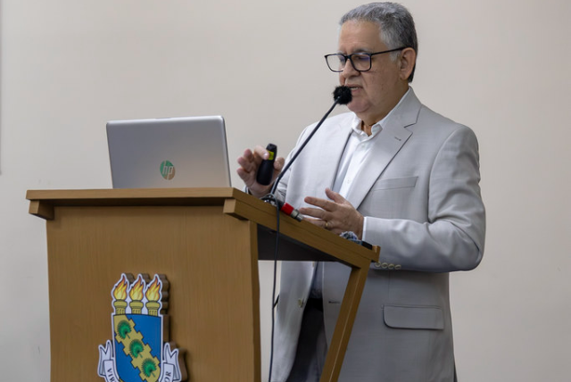 Um homem grisalho de terno cinza claro e óculos de grau fala em um púlpito 