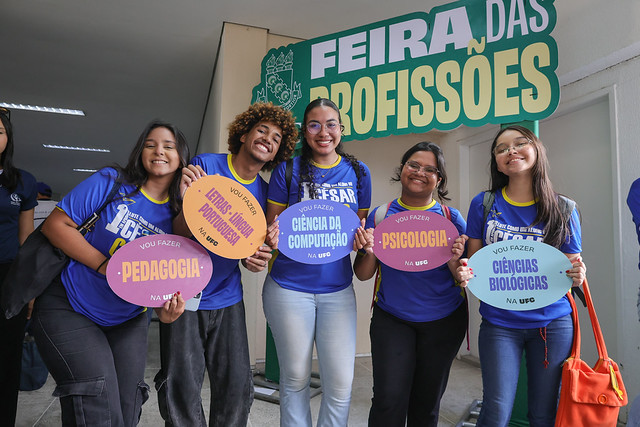 Imagem: cinco jovens estão de blusa azul e em pé. Elas seguram placas com nomes de cursos de graduação
