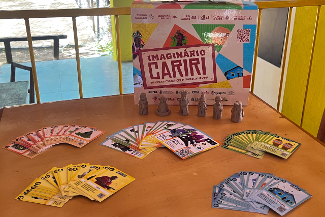 Imagem: Fotografia de uma mesa de madeira sobre a qual está exposto o jogo educativo “Imaginário Cariri”. Ao fundo, apoiada sobre a mesa, há uma caixa colorida do jogo com o título em destaque no centro. A embalagem apresenta ilustrações e elementos gráficos inspirados na cultura do Cariri cearense, além de logotipos institucionais e um código QR no canto superior direito. À frente da caixa, estão alinhadas cinco pequenas peças tridimensionais em tom amadeirado, representando personagens ou figuras do jogo. Sobre a mesa, diversos conjuntos de cartas coloridas encontram-se organizados em leques. À esquerda, há cartas em tons de rosa; no centro, cartas multicoloridas com ilustrações; à direita, cartas verdes. Na parte inferior da imagem, aparecem mais dois grupos de cartas: um conjunto amarelo à esquerda e outro azul-claro à direita. O ambiente parece ser uma área interna bem iluminada. Ao fundo, há uma grade metálica amarela e uma janela ou abertura que permite visualizar uma área externa com piso azul, árvores e um banco.