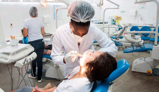Imagem: Profissional de odontologia, usando máscara, luvas e touca, realiza exame bucal em uma paciente reclinada em uma cadeira odontológica. Ao fundo, há outros consultórios e pessoas em atendimento em uma clínica odontológica.