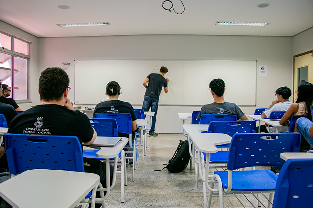 Imagem: estudantes de costas estão sentados em cadeiras azuis em uma sala de aula. Todos vestem blusa preta e estão olhando para um quadro branco, onde um professor está escrevendo