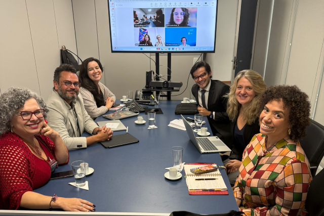 Seis pessoas estão sentadas ao redor de uma mesa azul olhando para a foto. Ao fundo, em uma tela de televisão, estão mais quatro pessoas participando virtualmente da reunião