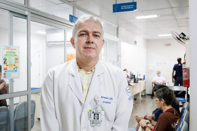 Imagem: Foto do médico geriatra e pesquisador Rômulo Lôbo utilizando jaleco branco e crachá no corredor do ambulatório de Geriatria do Hospital Universitário Walter Cantídio