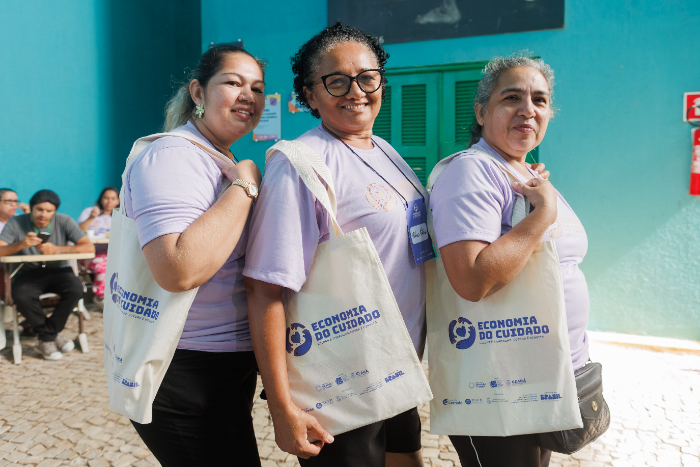 Três mulheres participantes do curso posam sorrindo para a foto em um espaço de atividades formativas. Vestindo camisetas lilás, elas carregam bolsas de tecido com a identidade visual do projeto “Economia do Cuidado”